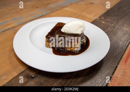 A white plate of delicious sticky toffee pudding chocolate cake with cream and chocolate sauce on a wooden kitchen work top Stock Photo