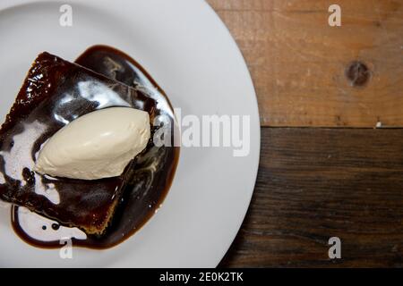 A white plate of delicious sticky toffee pudding chocolate cake with cream and chocolate sauce on a wooden kitchen work top Stock Photo