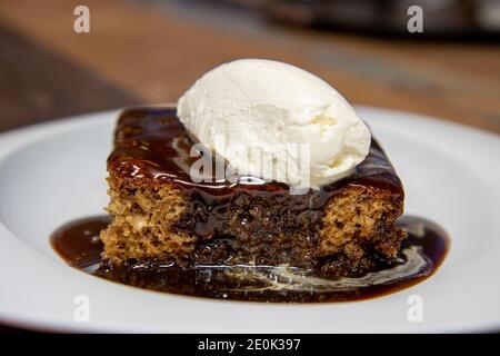 A white plate of delicious sticky toffee pudding chocolate cake with cream and chocolate sauce on a wooden kitchen work top Stock Photo