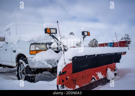 A white truck with a red snow plow and add on lights parked in the snow in front of a train-car home. Stock Photo