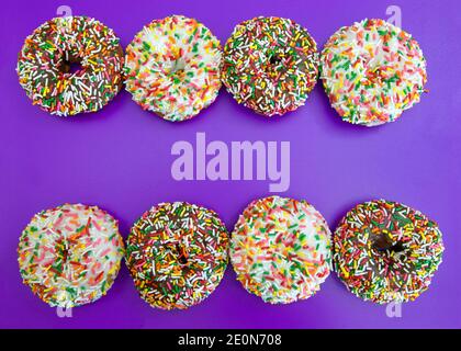 Flat lay view of many frosted donuts, chocolate and vanilla with candy sprinkles arranged in a row top and bottom on a purple background. Stock Photo
