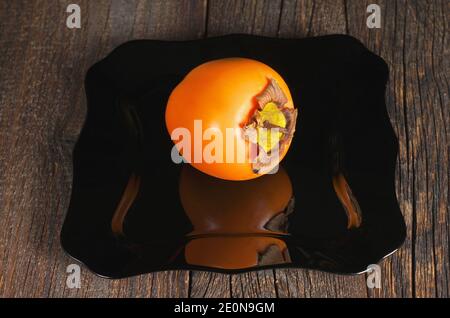 One persimmon fruit in black plate on dark wooden table Stock Photo