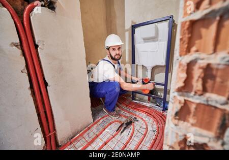 Plumber working in unfinished apartment, installing flush tank, looking at camera. Red pipes on the floor, heating system. Concept of building refurbishment, plumbing works and home renovation Stock Photo