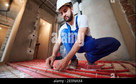 Horizontal, low angle view snapshot of young plumber wearing blue overalls and white helmet, tying up red tubes on the floor heating system in new unfinished apartment. Concept of home renovation. Stock Photo
