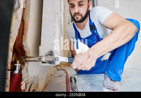 Horizontal snapshot of handsome plumber screwing plumbing fittings with wrench. Close-up of strong arm working with a tool during installation works in new apartment. Focus on hand with instrument. Stock Photo