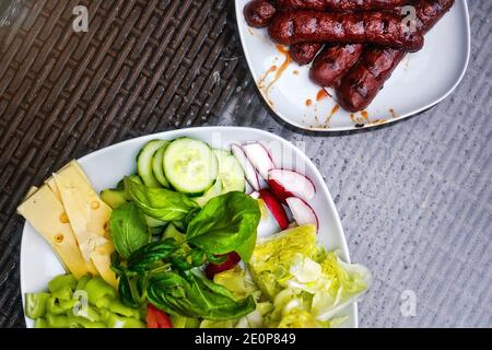 Two white plates, one with grilled bratwursts / frankfurters, other with salad - cucumbers, radish, basil, pepper and cheese. View from above Stock Photo