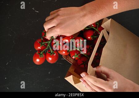 Taking out red cherry tomatoes with green leaves from brown paper bag over dark marble like board, detail on woman hand holding fruits Stock Photo