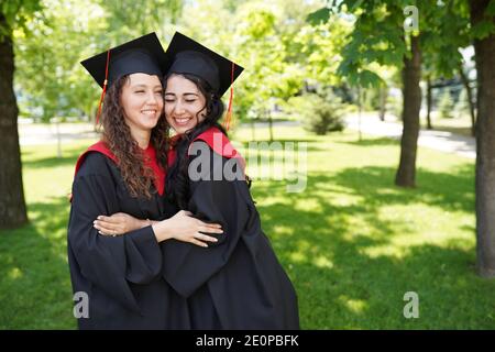 Successful graduates in academic dresses looking at camera and smiling outdoors in green park, place for text Stock Photo