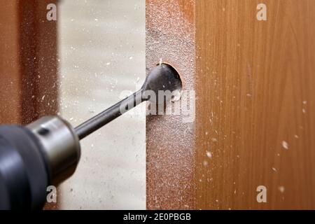 The locksmith uses a flat spade bit  for wood  to drill a handle hole with a lock in an interior wooden door. Stock Photo