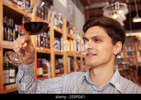 Close up of a cheerful young man tasting wine at wine cellar. Selective focus on red wine glass in the hand of a happy man Stock Photo