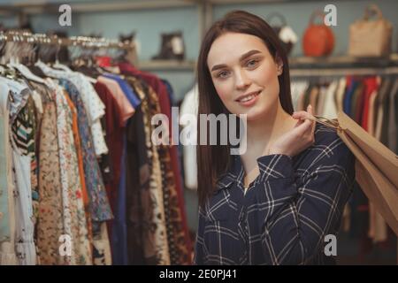 Beautiful dark haired young woman smiling to the camera, holding shopping bag, standing at clothing store. Charming female customer carrying her purch Stock Photo