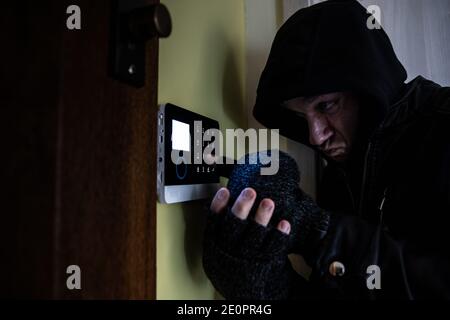 A burglar with gloves and a hood on his head tries to disarm the alarm system. Stock Photo