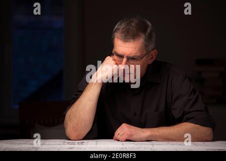 Pensive engineer or architect in dark shirt looking at blueprint at home by night - focus on the face Stock Photo