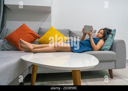 Asian woman lying on sofa and using tablet at home Stock Photo