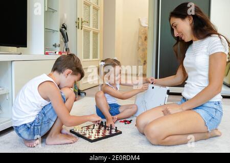 Little brother and sister playing chess at home with their mother Stock Photo