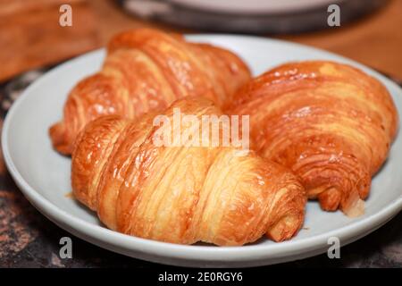 A plate of three freshly made croissants on a plate in a kitchen on a wooden work top Stock Photo