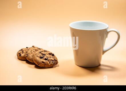 Close up white mug of milk and three dark chocolate chip cookies on empty background, copy space area. Yellow golden background Stock Photo