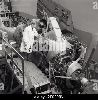 1950s, historical, wearing a white-coat and standing on a platform by the cockpit of an aircraft, a senior male engineer overseeing three young male aviation engineering trainees or apprentices, working on the engine of the training aircraft which has a propeller, England, UK. Stock Photo