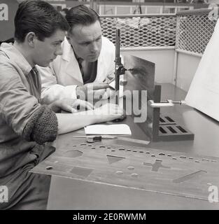 1950s, historical, a young aviation engineering trainee or apprentice sitting with his tutor, a senior engineer, being shown how to use a sharp tool or blade to cut different geometric patterns on a metal sheet. Geometry, a branch of mathematics, is all about shapes and their properties and its knowledge is important for work in the areospace sector. Stock Photo