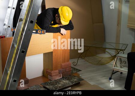 An employee leans against the cupboard and looks down at the glued brick-shaped decorative tiles. An employee dressed in all personal protective equipment. Stock Photo