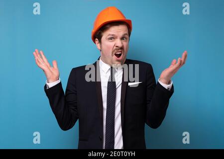Caucasian young man in orange protective helmet asking what feeling upset. Studio shot on blue wall. Stock Photo