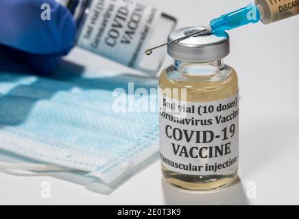 Covid-19 coronavirus vaccine with hypodermic syringe needle with droplet of liquid on tip. Gloved hand holding bottle in background Stock Photo