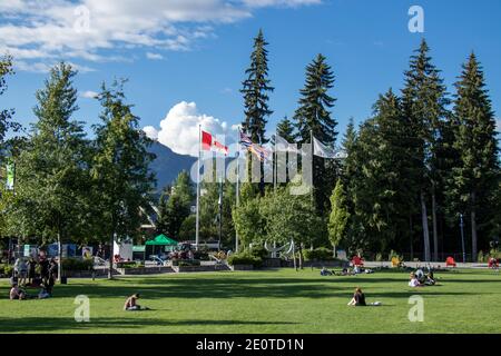 Whistler, Canada - July 5,2020: View of poles with waving flags in the Whistler Village. People are sitting on the green lawn on a sunny day Stock Photo