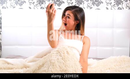 female rises in bed with an alarm clock in her hands in horror realizes that she slept. Concept of lack of time, being late for work or school Stock Photo