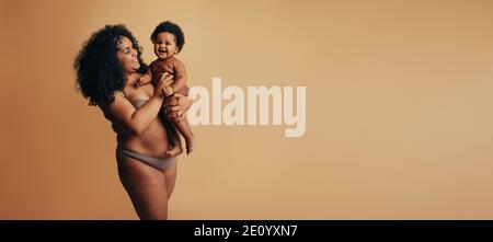 African american woman carrying her child in her arms over brown background. Cheerful mother and baby. Stock Photo