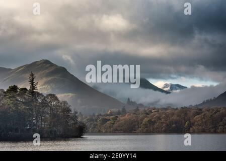Epic landscape image looking across Derwentwater in Lake District towards Catbells snowcapped mountain with thick fog rolling through valley