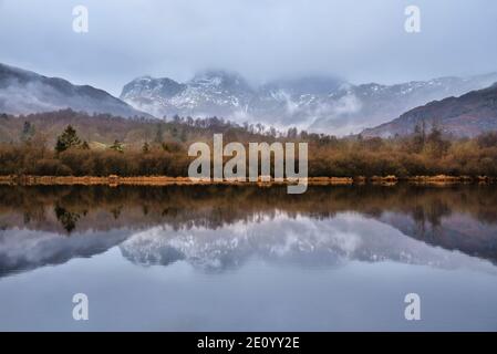Epic dramatic landscape image looking across River Brathay in Lake District towards Langdale Piks mountain range on mistry Winter morning Stock Photo