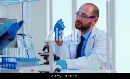 Doctor dictating to nurse virus results informations looking on test tubes in modern equipped laboratory. Doctors examining vaccine evolution using high tech researching diagnosis against covid19 Stock Photo