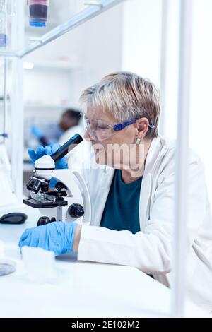 Concentrated senior scientist conducting genetic experiment using microscope. Elderly researcher carrying out scientific research in a sterile lab using a modern technology. Stock Photo