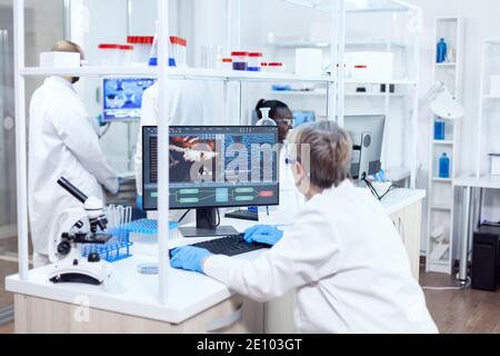 Elderly researcher engineering human adn using softtware on powerfull computer. Senior scientist in pharmaceuticals laboratory doing genetic research wearing lab coat with team in the background. Stock Photo