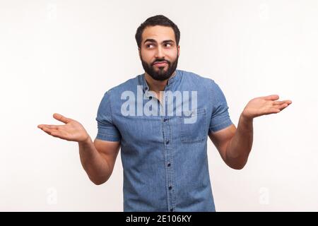 Puzzled clueless man with beard in blue shirt throwing up his hands looking at camera with uncertain expression, has no answers. Indoor studio shot is Stock Photo