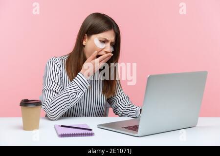 Tired sleepy woman yawning closing mouth with hand, working on laptop sitting at home office and wearing eyes patches. Indoor studio shot isolated on Stock Photo
