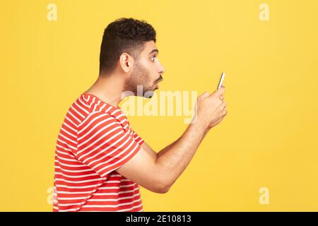 Profile portrait surprised shocked man with beard in red striped t-shirt looking at smartphone display with astonishment, confused with tariffs. Indoo Stock Photo