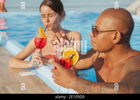 Handsome African man and his beautiful girlfriend enjoying cocktails in the swimming pool at resort hotel. Happy couple having drinks at the poolside. Stock Photo