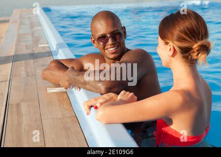 Attractive cheerful African man talking to his girlfriend at the swimming pool. Multiethnic couple enjoying summer vacation together, resting after sw Stock Photo