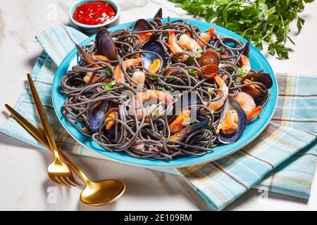 Black squid ink pasta with seafood: mussels and shrimps with white wine and garlic sauce on a blue plate with golden cutlery on a white marble backgro Stock Photo