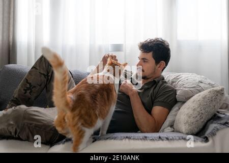 young man lying on the sofa under the window plays with a brown and white cat, Stock Photo