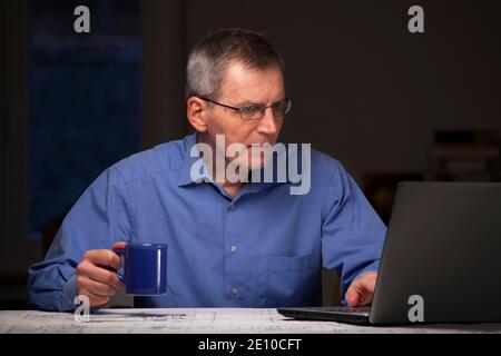 Mature businessman or manager working at home with laptop and a cup of coffee in his hand - dark background Stock Photo