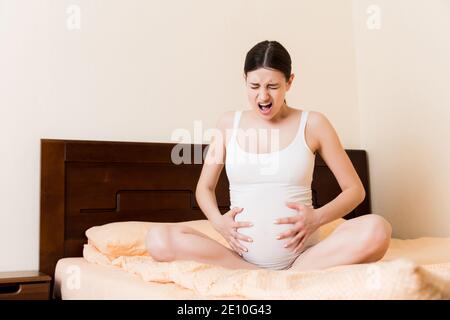 Young pretty pregnant woman, in pain, sitting on bed in bedroom, preparing to give a birth. Stock Photo