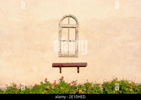 The rustic wooden window frame and the brown shelf below hang on the empty house wall. Below a flowerbed with blooming pink flowers. Stock Photo