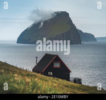 Black house on famous faroese Witches Finger Trail and Koltur island on background. Sandavagur village, Vagar island, Faroe islands, Denmark Stock Photo