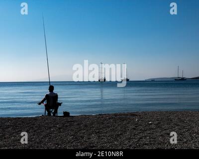 A lonely fisherman silhouette sits in a chair at the beach and tries to catch some fishes. A cat lies beside him and waiting for food. Sailing boats Stock Photo