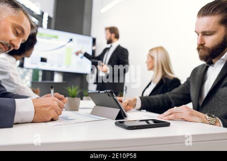 Two focused diverse businessmen, making notes while listening to the speech of male speaker, showing financial presentation to diverse employees group Stock Photo