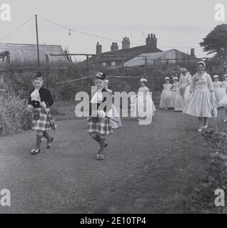 1950s, historical, two young boys in kilts leading the Rose Queen and her entourage of young girls in their gowns through a local park, to where the ceremony will take place, Farnworth, Lancashire, England, UK.  Rose Queen day was a special day for the children of the town who attended the church's Sunday School and was connected to the traditional 'summer solstice', which had its origins in the old pagan festivals. It was sometimes done together with the walking Day when the children would gather and walk the streets. Stock Photo