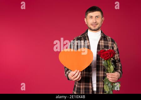 Portrait of a young man with a red paper heart in his hands and a bouquet of red roses. St.Valentine's Day. Red background with empty space. High qual Stock Photo