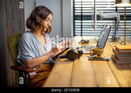 Young woman dressed casually having some creative work, drawing on a digital tablet, sitting at the cozy and stylish home office Stock Photo